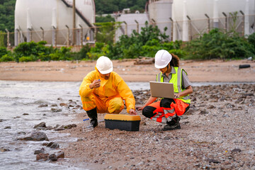 Two environmental engineers in safety gear work at a shoreline near gas storage tanks. One collects...