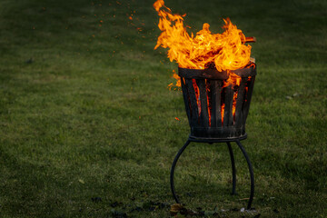 A metal fire basket with tall, bright orange flames burns on a grassy lawn, with embers rising into...