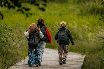 Three children with backpacks walk together along a wooden path through lush greenery, heading away into a peaceful natural landscape.
