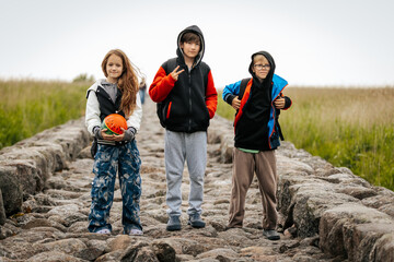 Three children stand on a stone path in nature, smiling and posing casually. One holds a ball, and the others wear hooded jackets and backpacks.