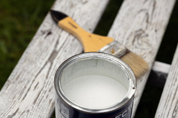 A can of white paint sits on a weathered wooden bench. A paintbrush rests beside it, ready for use....
