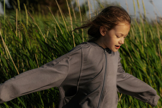 A young girl walks playfully along a sandy beach with wind blowing her hair, dressed in a gray hoodie and patterned pants, near tall green grass. - Powered by Adobe