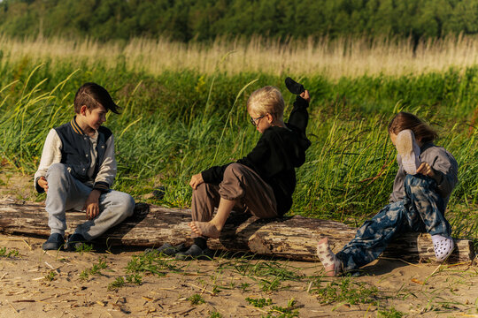 Three children sit and play on a driftwood log at the edge of a grassy dune, enjoying a sunny and breezy day in nature.