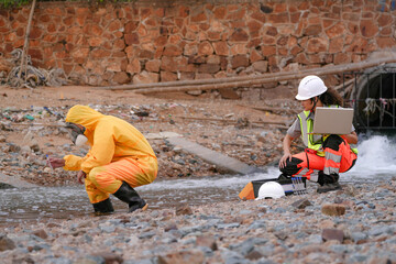 Environmental field team in protective gear analyzes water samples on a polluted beach. One holds a laptop while the other handles vials for chemical testing of coastal pollution.