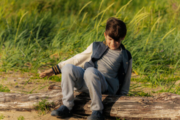 A young boy in casual clothes sits on a large weathered log in a grassy sandy area, smiling and looking down as sunlight illuminates the scene.