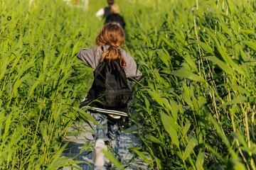 Children with backpacks walk through a dense field of tall green grass along a narrow wooden path,...