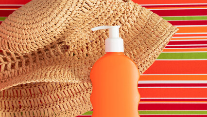 A bottle of sunscreen and a hat lie on a striped lounge chair. Summer beach concept. Orange tube of sunscreen close-up.