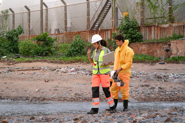 Two environmental researchers walk back from shoreline testing. They carry safety gear, a toolbox, a respirator mask, and a laptop after collecting water samples from a contaminated coast.