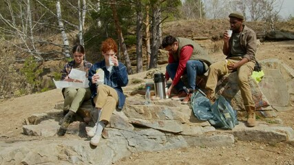 Biracial group of volunteers sitting on rock, communicating, laughing and enjoying food after long cleanup day