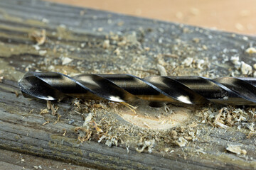 A close-up of a wood drill bit resting on a wooden surface with wood shavings scattered around. The...
