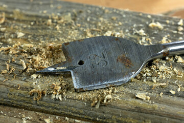 A wooden drill bit rests on a wooden surface covered with wood shavings. The drill bit is metallic...