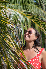 A beautiful young woman wearing a red swimsuit and sunglasses smiles as she enjoys the tropical...