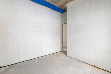 The room is under construction with a rough finish, plastered walls, concrete floor. Wiring of ventilation pipes on a concrete ceiling in a house under construction. Living Room Bedroom Area