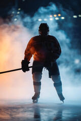 Obraz premium Dark silhouette of a male hockey player in a uniform, helmet and skates with a stick on the ice arena with smoke and blue back light. Sportsman posing at the stadium ice rink. Winter sports.