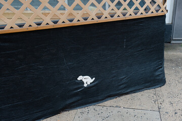 Small dog figure peeking out from under a black tarpaulin in an urban setting on a sunny day