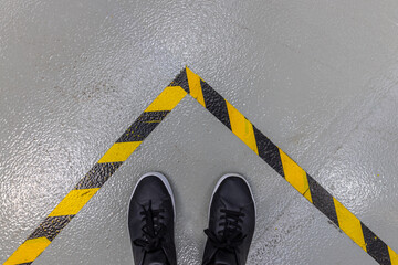 A person's feet in black shoes stand on a grey concrete floor, bordered by a prominent yellow and...