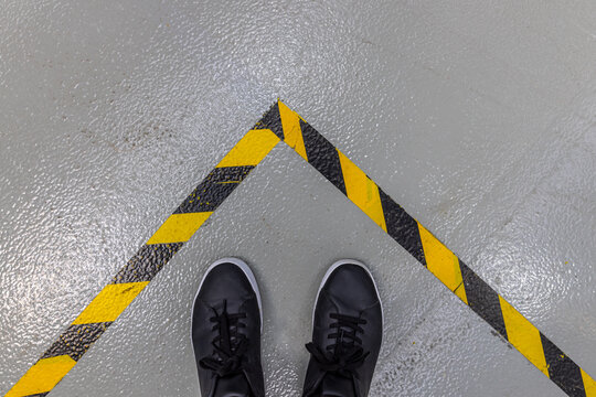 A person's feet in black shoes stand on a grey concrete floor, bordered by a prominent yellow and black striped hazard warning line. - Powered by Adobe