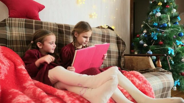 Two little sisters snuggling up on the sofa in a cozy living room at Christmas at home during winter break. Kids reading a book.