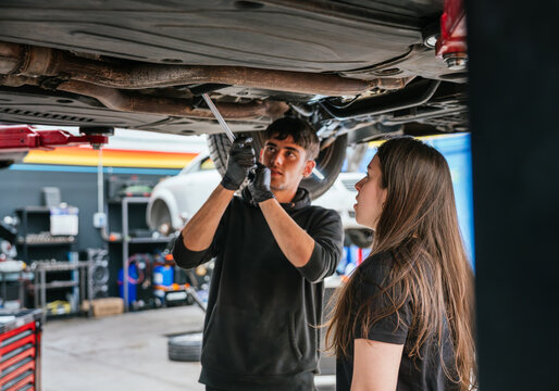 Young mechanic woman watches attentively as a colleague works under a lifted vehicle in a car workshop