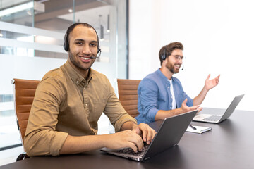Two diverse customer service representatives wearing headsets are working on laptops in a modern office environment.