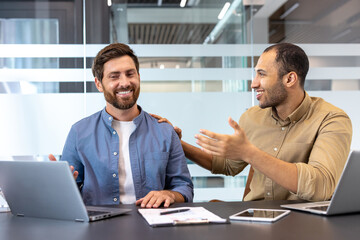 Two diverse male colleagues smile and collaborate in a modern office setting, using laptops and...