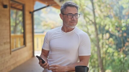 Mature man enjoying peaceful morning in wooden cabin. Businessman drinking coffee on balcony of mountain chalet. Entrepreneur working with smartphone. Relaxation and getaway, ideal lifestyle. - Powered by Adobe