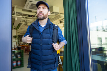 Man with delivery bag exiting cafe in blue vest and cap