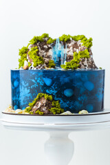 Beautiful birthday cake in the shape of an island decorated with chocolate edible mountains, greenery with waterfall. Tropical island cake surrounded with blue jelly ocean water