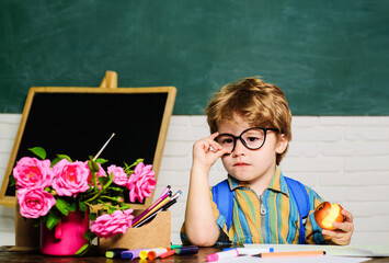 School break. Lunch time. Cute serious schoolboy sitting at table and eating apple in classroom....