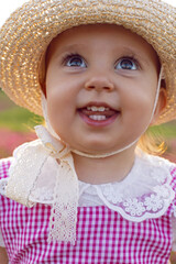 Beautiful one-year-old baby girl stands in a pink dress and a straw hat in a field at sunset among bright flowers.