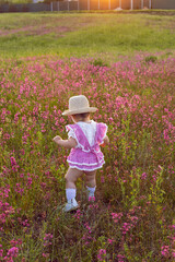 Beautiful one-year-old baby girl stands in a pink dress and a straw hat in a field at sunset among bright flowers.