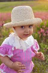 Beautiful one-year-old baby girl stands in a pink dress and a straw hat in a field at sunset among bright flowers.