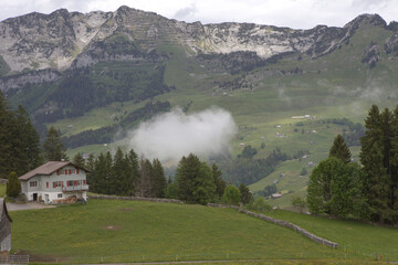 Breathtaking alpine landscape in Switzerland with green rolling hills, low-hanging clouds, and...