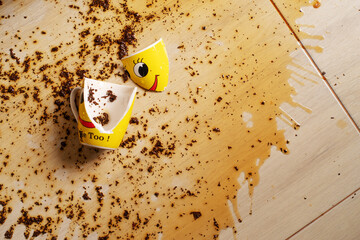 Broken yellow cup with smiley face and spilled black tea on the floor. Tea spilled on the floor. Splashes and tea leaves on the floor.