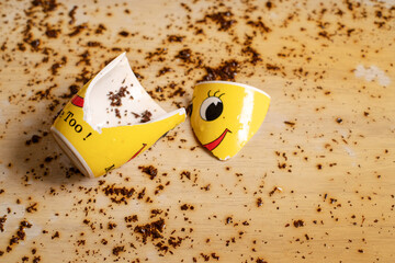 Broken yellow cup with smiley face and spilled black tea on the floor. Tea spilled on the floor. Splashes and tea leaves on the floor.