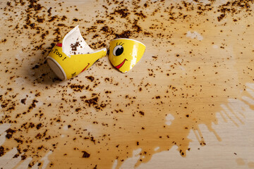 Broken yellow cup with smiley face and spilled black tea on the floor. Tea spilled on the floor. Splashes and tea leaves on the floor.