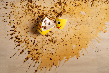 Broken yellow cup with smiley face and spilled black tea on the floor. Tea spilled on the floor. Splashes and tea leaves on the floor.