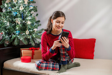 Smiling woman in red pajamas using mobile phone near Christmas tree and gifts. Happy holiday season morning concept.