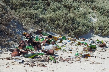 Waste contamination polluting a pristine sandy beach with rusty cans, broken bottles, and other...