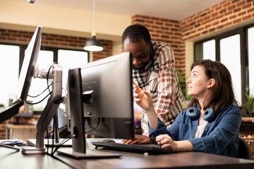 Black male manager standing by desk, discussing product designs with caucasian employee working on...