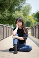 A young teenager sits comfortably on a sunlit wooden bridge, casually dressed in a black top, jeans, and black boots, trees lining the background.