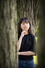 A young teenager stands pensively among tree trunks, framed by bark and soft natural light, wearing a black top and jeans in a calm forest scene.
