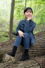A young teenage girl in denim sits calmly on a fallen tree in a lush green forest, gazing off thoughtfully, surrounded by greenery.