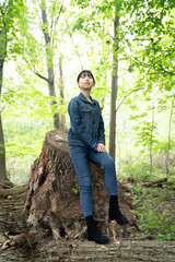 A young teenage girl in denim sits calmly on a fallen tree in a lush green forest, gazing off thoughtfully, surrounded by greenery.