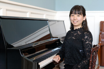A young teenage girl in a black blouse sits gracefully by a grand piano, smiling at the camera in a bright, elegant room.