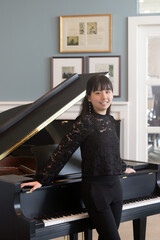 A young teenage girl in a black blouse stands gracefully by a grand piano, smiling at the camera in a bright, elegant room.