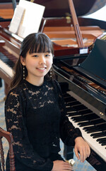 A young teenage girl in a black blouse stands gracefully by a grand piano, smiling at the camera in a bright, elegant room.