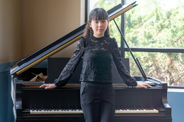 A young teenage girl in a black blouse stands gracefully by a grand piano, looking at the camera in a bright, elegant room.