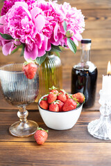 A glass of dark wine decorated with strawberries, Pink peony flowers in a vase and a bottle of red wine