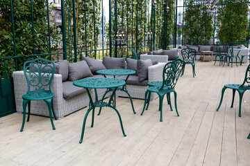 A modern grey sofa with cushions is installed in a glass greenhouse outside. Lush green plants and vibrant flowers surround the living area, creating a serene atmosphere.